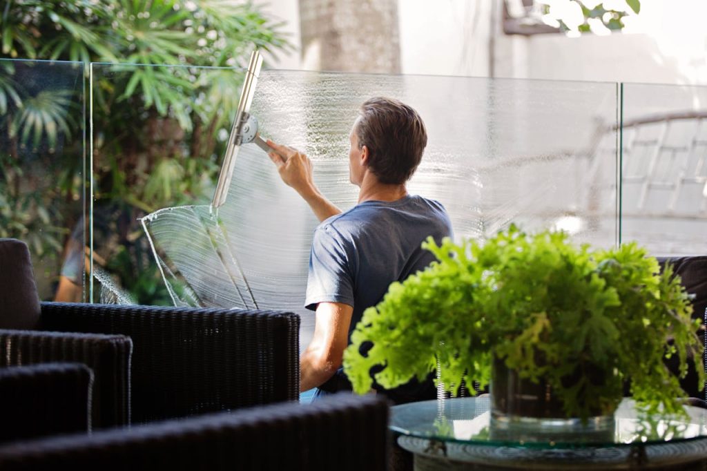 man-in-gray-shirt-cleaning-clear-glass-wall-near-sofa-713297 A man cleaning a glass barrier outdoors with a squeegee, surrounded by greenery.